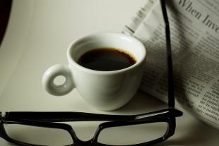 Glasses and a cup of coffee resting next to a newspaper on a white table 